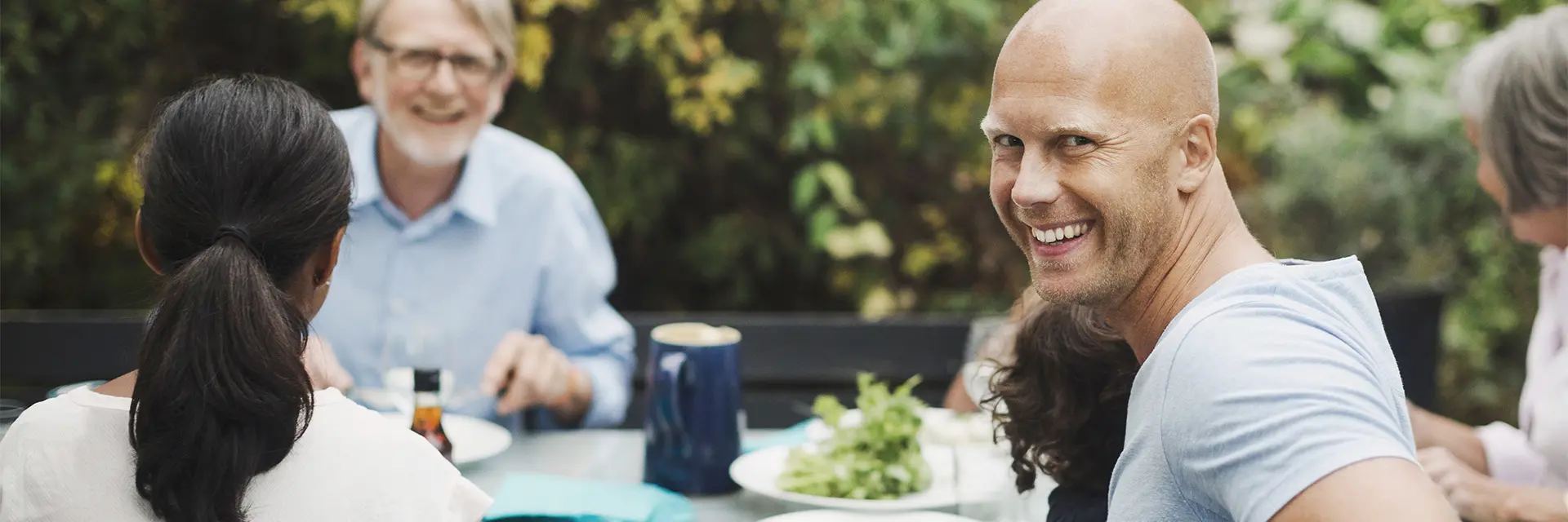 Familiemiddag ute på sommeren, tre generasjoner, mann ser mot oss og smiler.