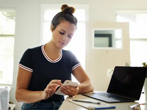 Kvinne sitter foran en datamaskin og notatblokk med telefonen i hånda og ser tankefull ut