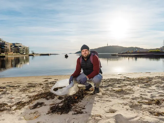 Mann med søppelpose i hendene sitter på huk på sandstrand med havet bak seg.