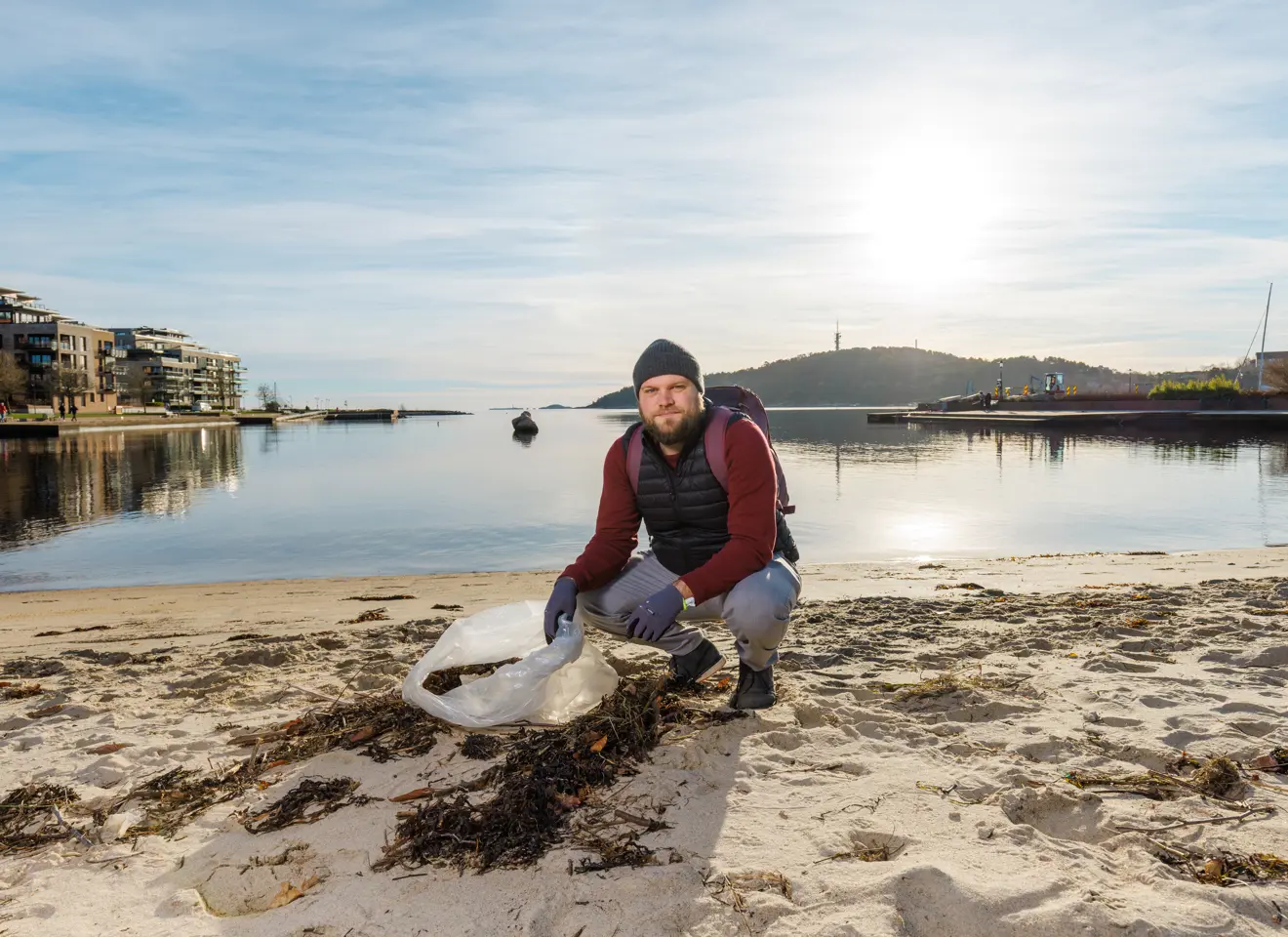 Mann med søppelpose i hendene sitter på huk på sandstrand med havet bak seg.