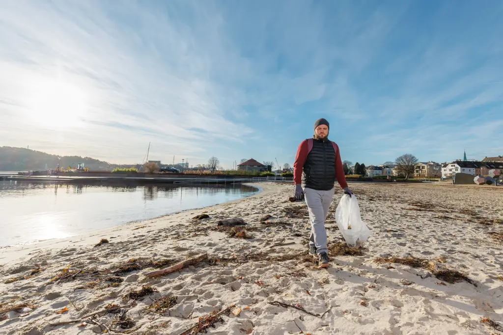 Mann går på stranda i Kristiansand og plukker søppel, det er sol, men kjølig vinter.