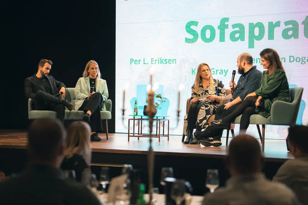 Tre kvinner og to menn sitter i lenestoler på en scene og diskuterer, en holder en mikrofon. En lysende storskjerm fyller veggen bak dem. 