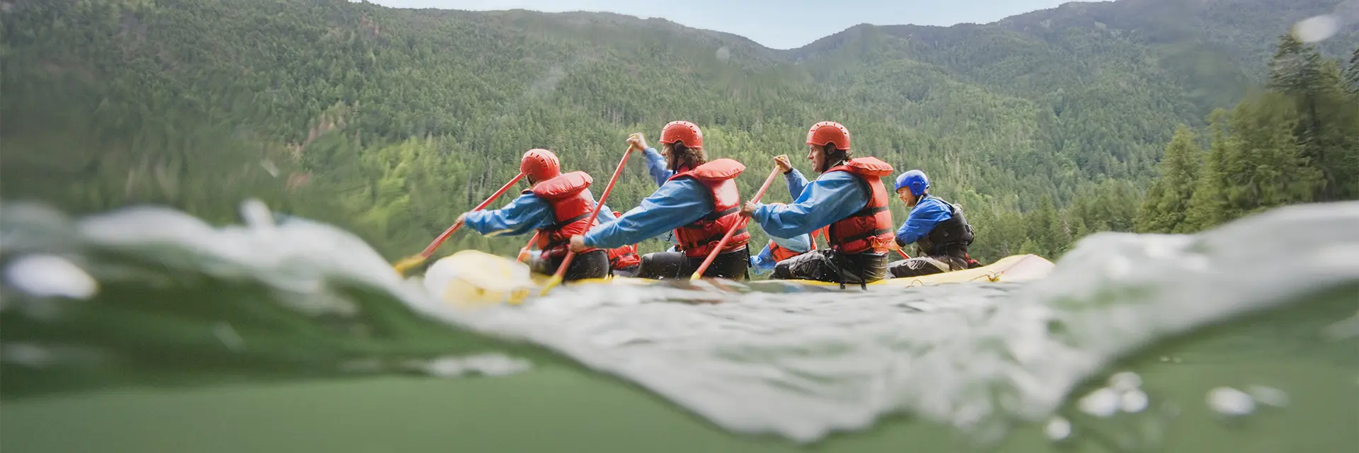 Full raftingbåt på vei nedover en elv, vann skvulper rundt dem, pent vær.