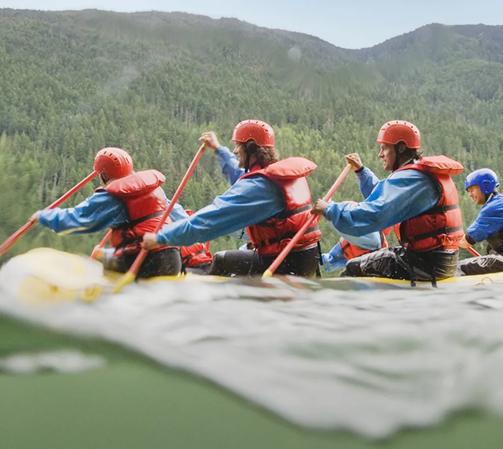 Full raftingbåt på vei nedover en elv, vann skvulper rundt dem, pent vær.