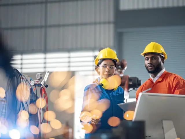 Mann og kvinne med hjelm i fabrikk, gnister i forgrunnen