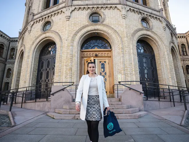 President Elisabet Haugsbø går foran inngangen til Stortinget, hvit jakke og mønstret skjørt, nett med Tekna-logo i hånda 