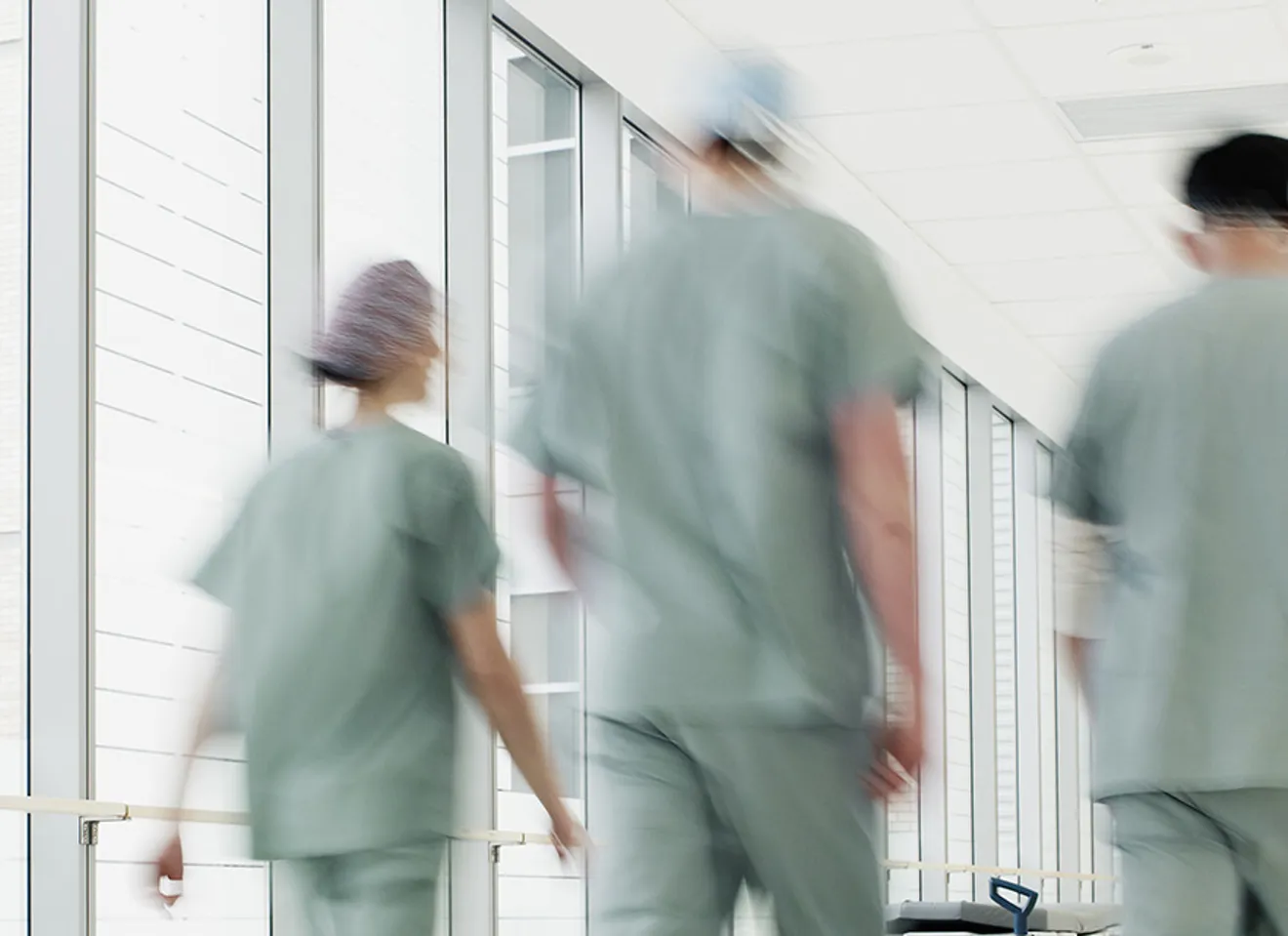 Interiør fra et sykehus, tre helsepersonell vandrer i en lang gang i grønne klær med ryggen til.