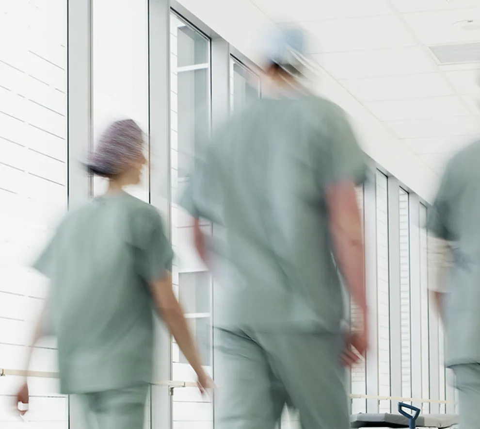 Interiør fra et sykehus, tre helsepersonell vandrer i en lang gang i grønne klær med ryggen til.