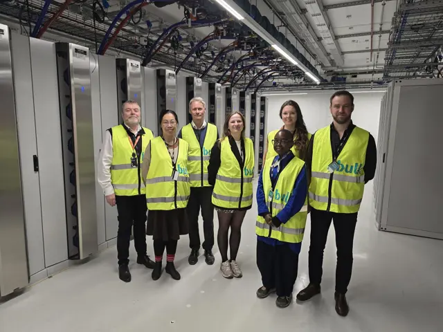 Gruppe på sju personer iført gule refleksvester står i en serverhall med rekker av serverrack og synlige kabler i taket.