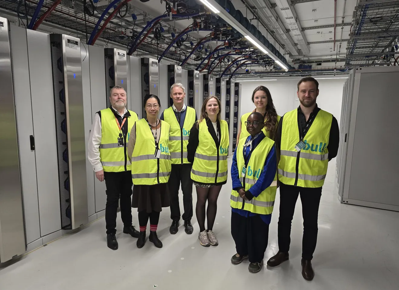 Gruppe på sju personer iført gule refleksvester står i en serverhall med rekker av serverrack og synlige kabler i taket.