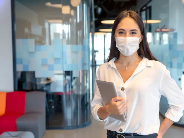 Illustrasjonsbilde av en ung kvinne med munnbind og nettbrett står i et tomt kontoret