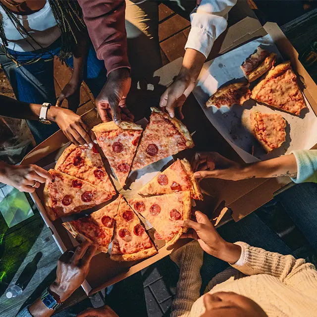 En gruppe mennesker samlet rundt et bord utendørs om kvelden for å dele pizza fra esker. Bildet er tatt i fugleperspektiv og viser hender som tar pizzastykker, samt flasker med drikke på bordet.
