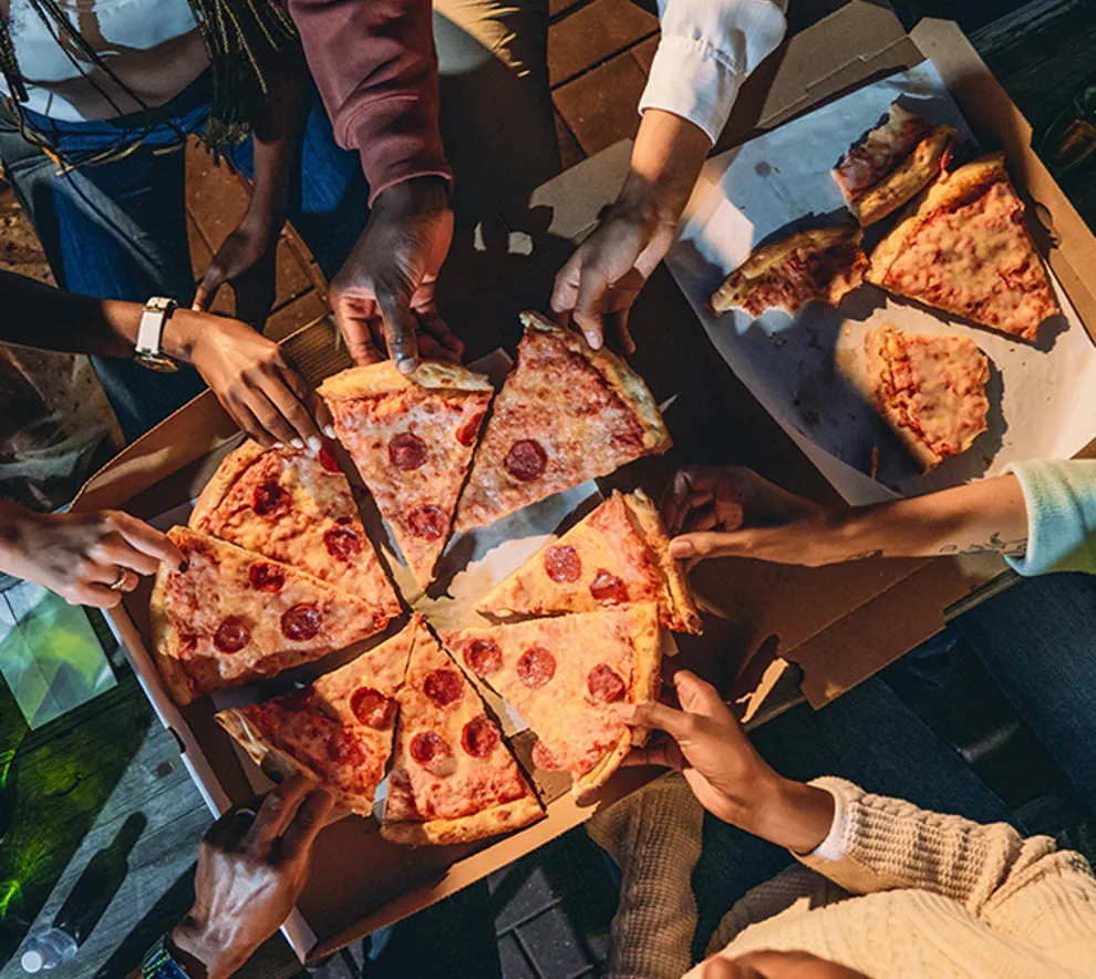 En gruppe mennesker samlet rundt et bord utendørs om kvelden for å dele pizza fra esker. Bildet er tatt i fugleperspektiv og viser hender som tar pizzastykker, samt flasker med drikke på bordet.