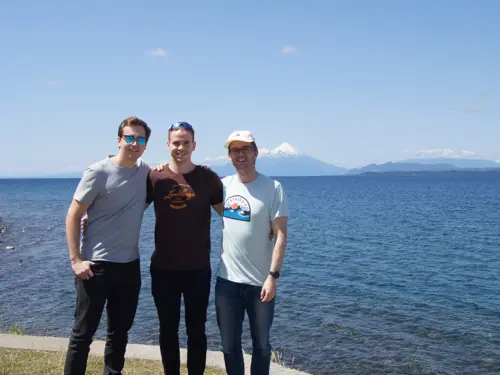 Tre unge menn står foran tropisk hav og smiler til kamera