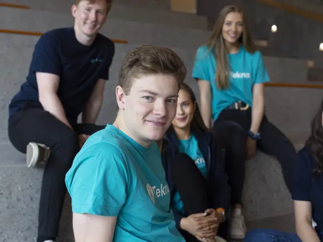 Person i turkis Tekna-tskjorte ser inn i kameraet og flere andre unge studenter sitter rundt ham i en steintrapp