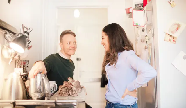 en ung mann og ung kvinne på et lite kjøkken med bakst og vannkoker på kjøkkenbenken
