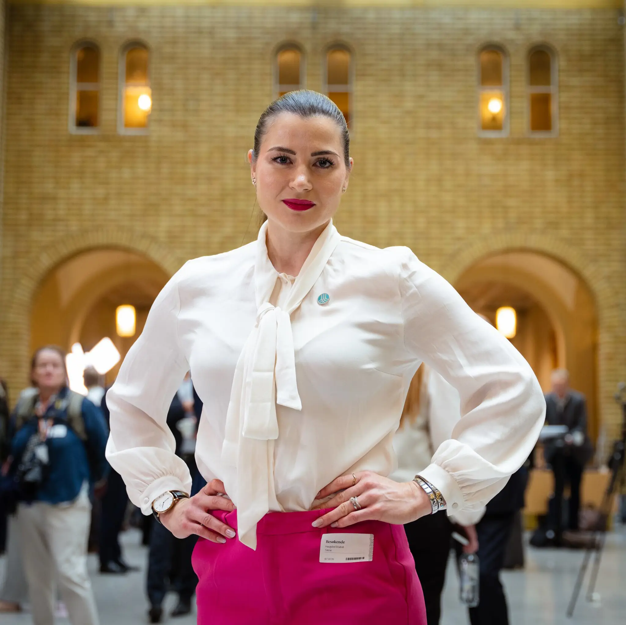 Elisabet haugsbø i vandrehallen på Stortinget, hvit bluse, rosa bukse, med armene i siden, bestemt ansiktsuttrykk