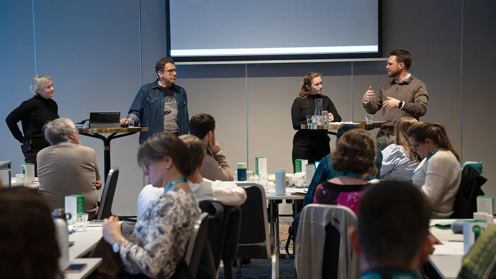 Fire personer i debatt i et stort rom, publikum ses bakfra 