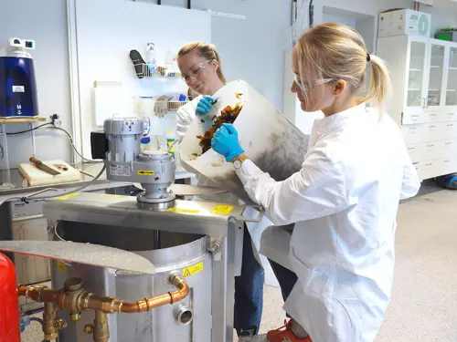 Weløy Aarseth og Sandberg Birgersson i laborfrakk og verneutstyr fyller sukkertare i en maskin