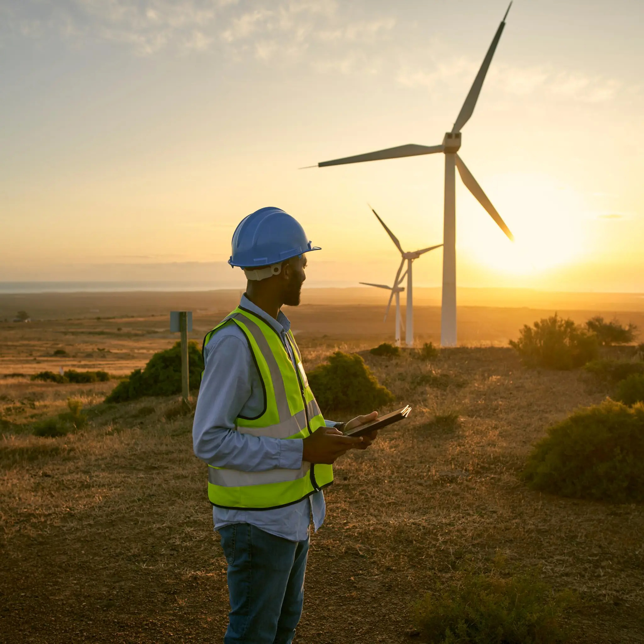 mann med refleks og nettbrett i hånda, ser mot vindmøller i solnedgang