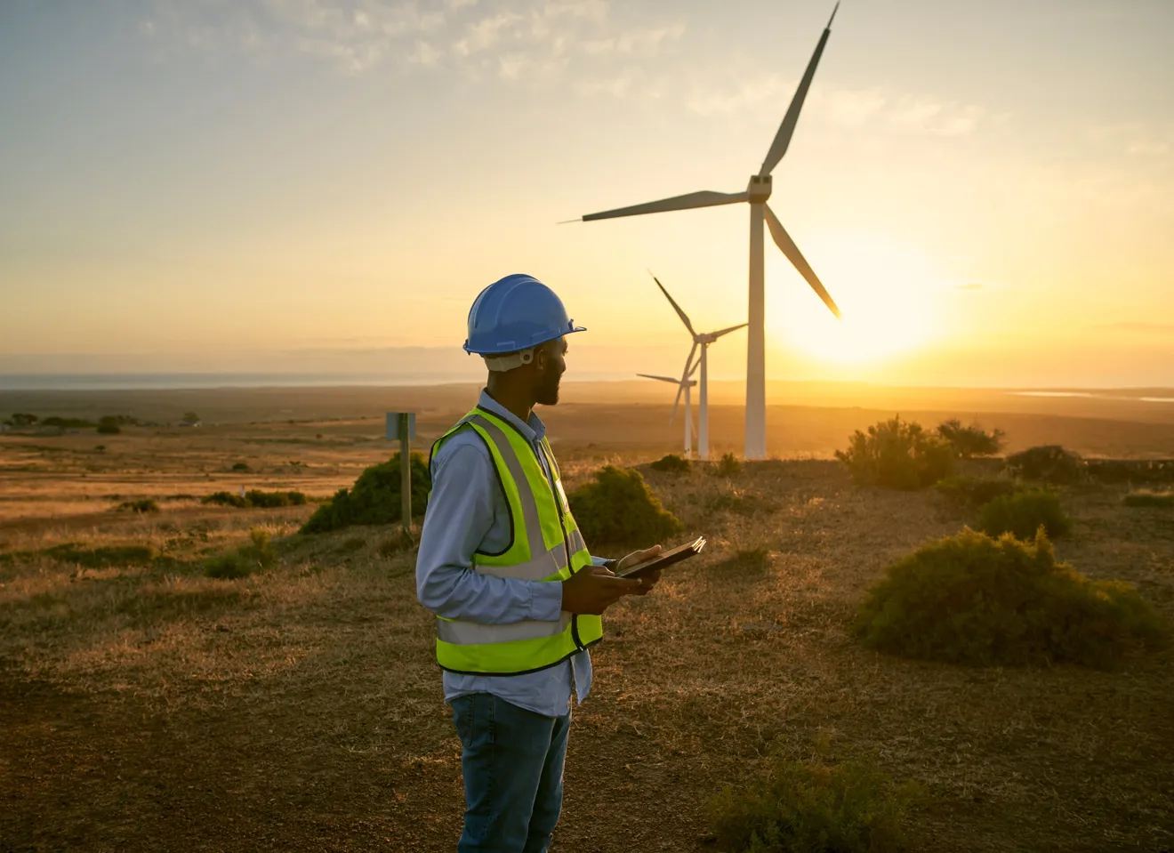 mann med refleks og nettbrett i hånda, ser mot vindmøller i solnedgang