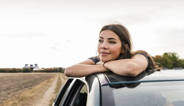 Kvinne som lener seg ut av solvinduet på en bil og ser ut i horisonten med et smil