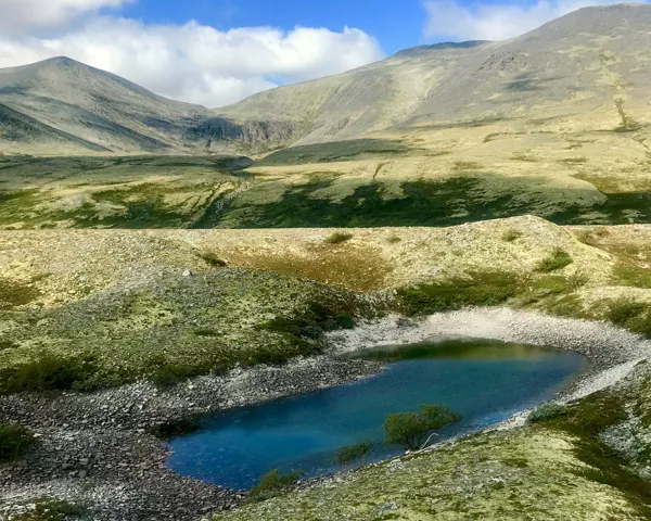Landskapsbilde av et lite vann omgitt av jord med veldig lite planter og fjell i bakgrunn
