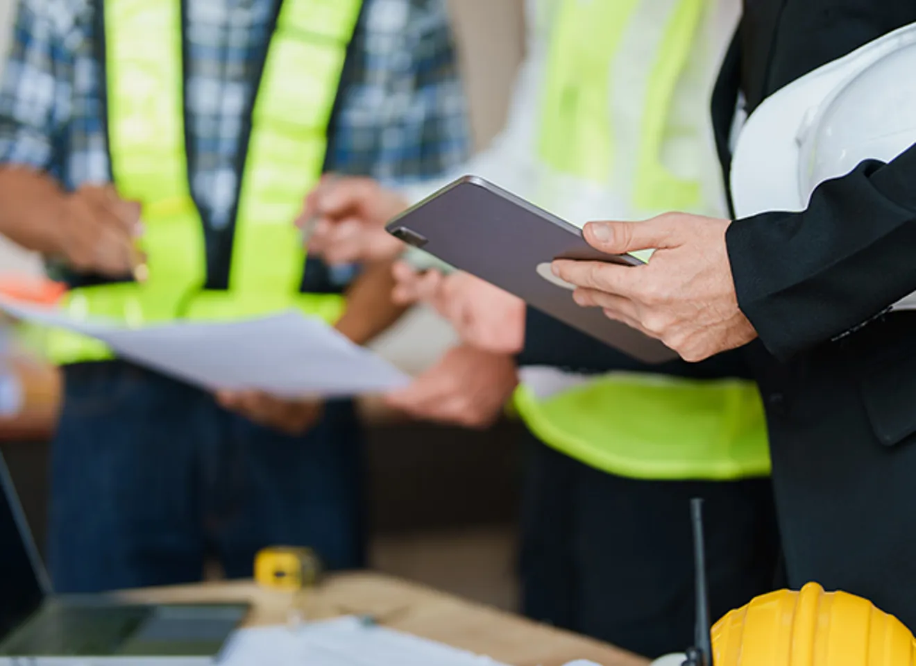 Gruppe av fagpersoner, inkludert en person i dress og arbeidere i sikkerhetsvester, som samarbeider om byggeplaner og dokumenter ved et bord.