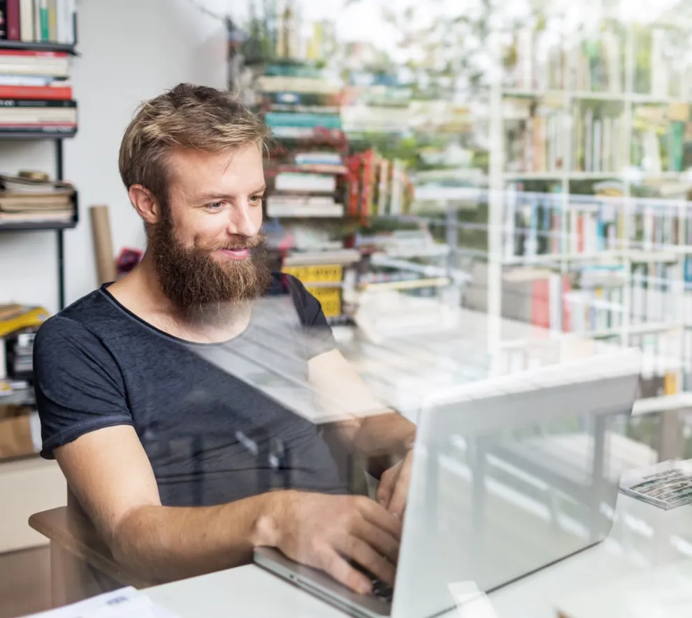 Ung mann med skjegg sitter foran en bærbar datamaskin på et hjemmekontor