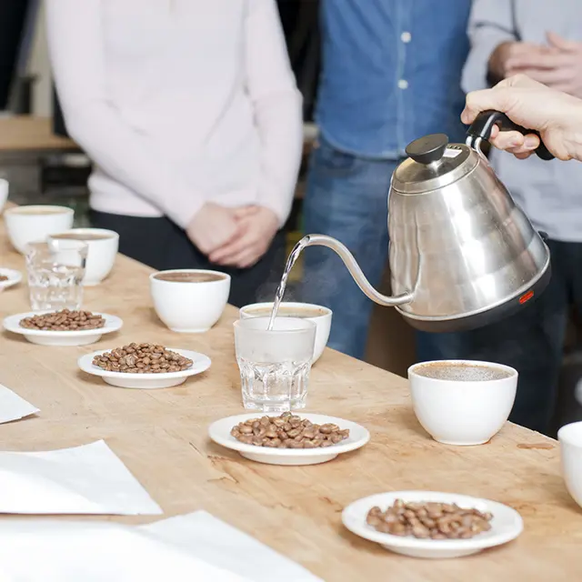 Person heller varmt vann fra en metallkjele over kaffekopper under en kaffesmaking. Kaffebønner er lagt frem på tallerkener. Fokus på kaffeprosess og smaking.