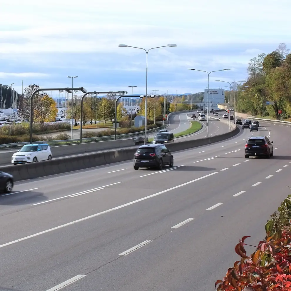 Biltrafikk på E18 med flere kjørefelt og veianlegg i urbane omgivelser.