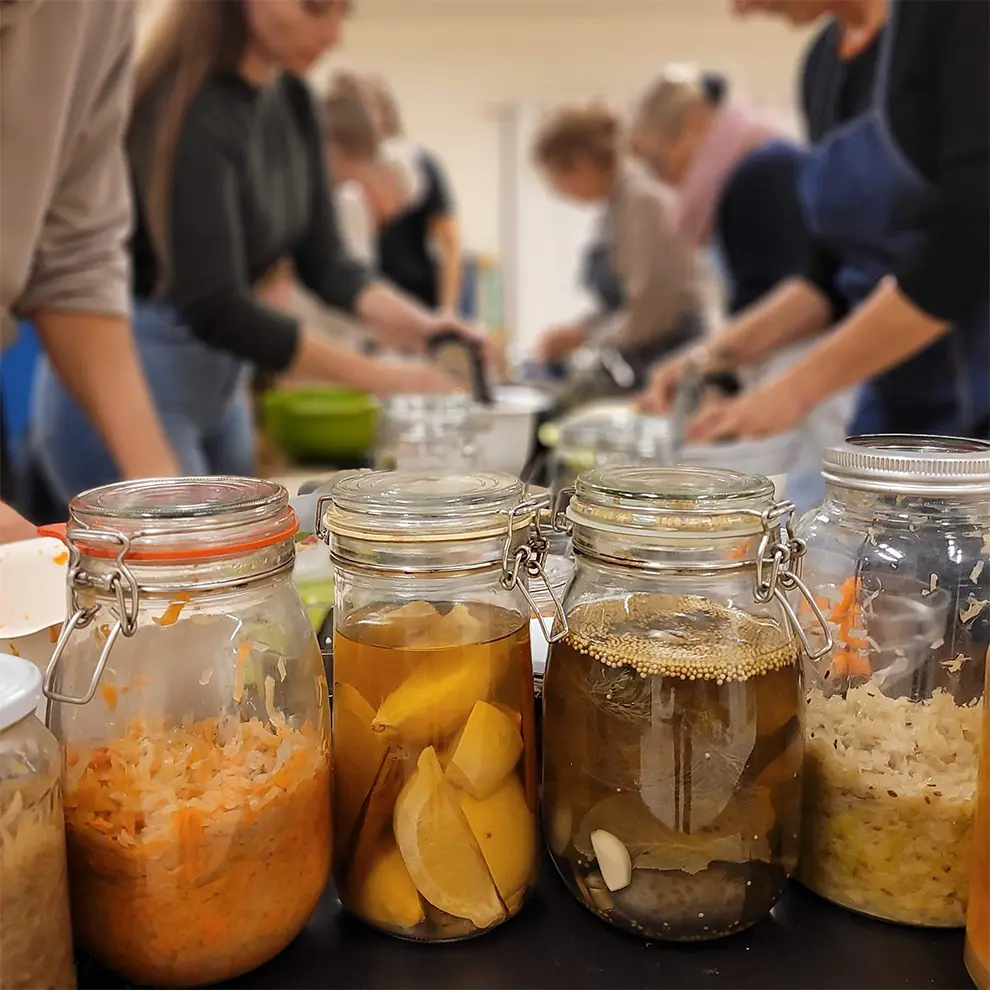 Norgesglass med variert innhold i fermenteringsprosessen, i bakgrunnen personer som forbereder matvarer til fermentering.
