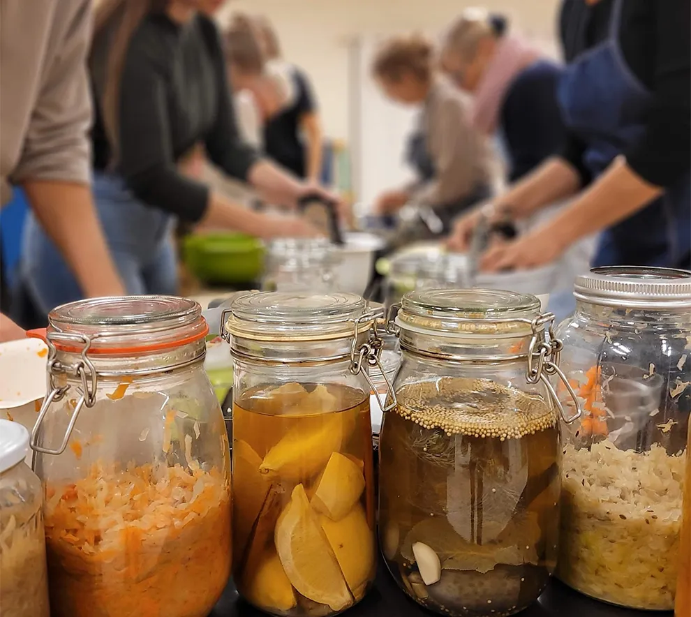Norgesglass med variert innhold i fermenteringsprosessen, i bakgrunnen personer som forbereder matvarer til fermentering.