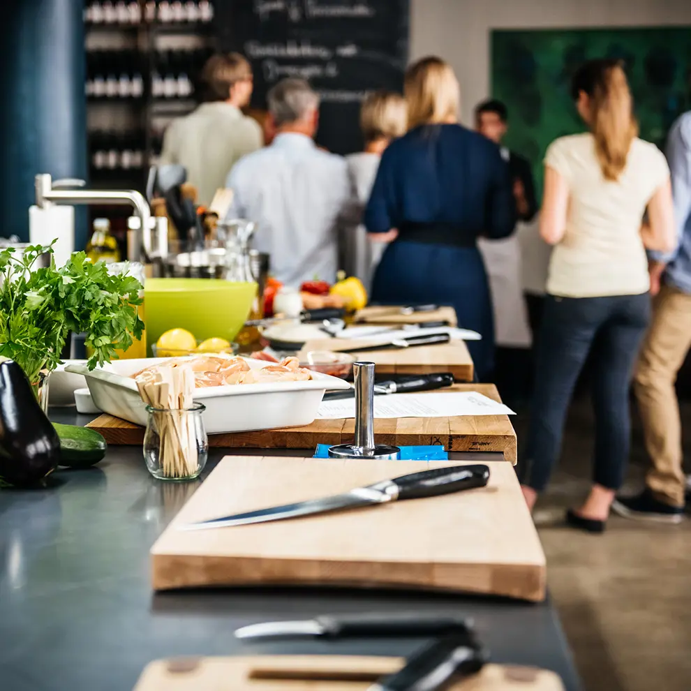 Matlagingskurs på profesjonelt kjøkken med råvarer, skjærebrett og deltakere i bakgrunnen.