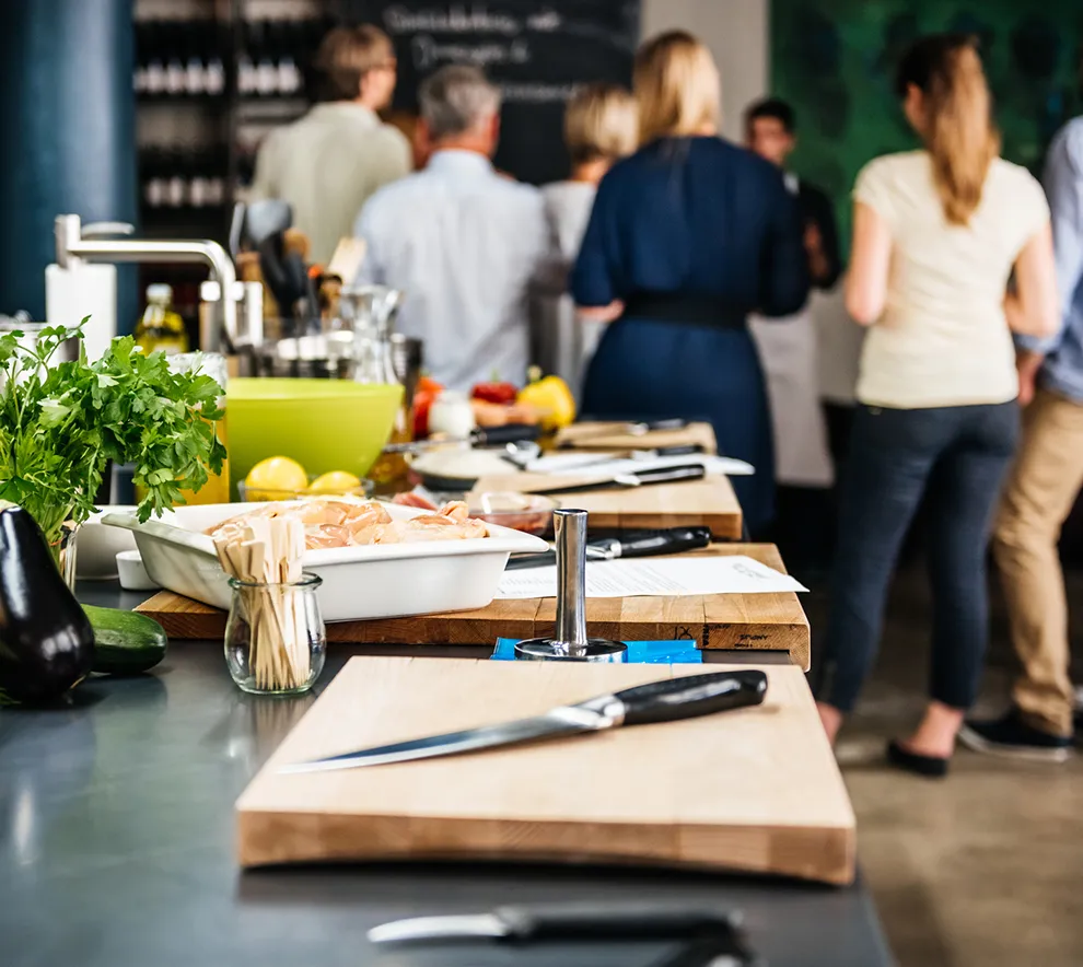 Matlagingskurs på profesjonelt kjøkken med råvarer, skjærebrett og deltakere i bakgrunnen.