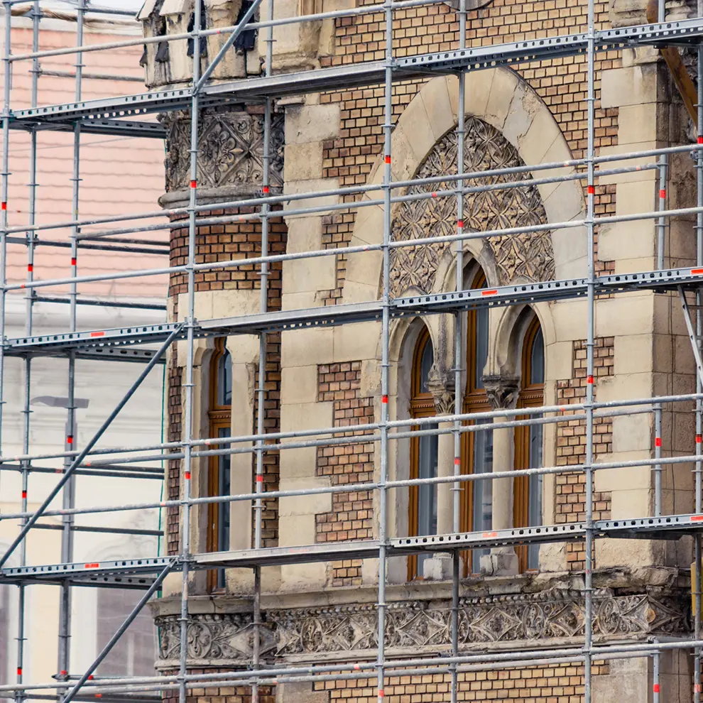 Stillaser rundt vernet murbygg under restaurering og ombruk av historisk fasade med buede vinduer og detaljer.