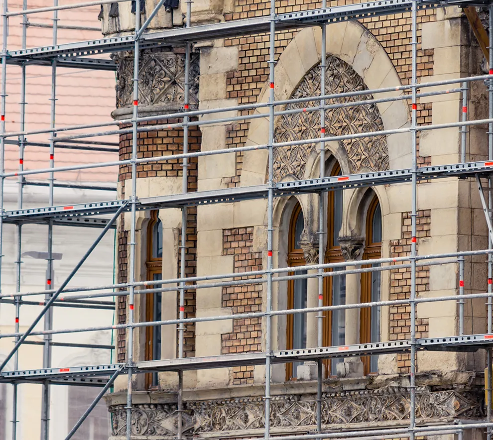 Stillaser rundt vernet murbygg under restaurering og ombruk av historisk fasade med buede vinduer og detaljer.