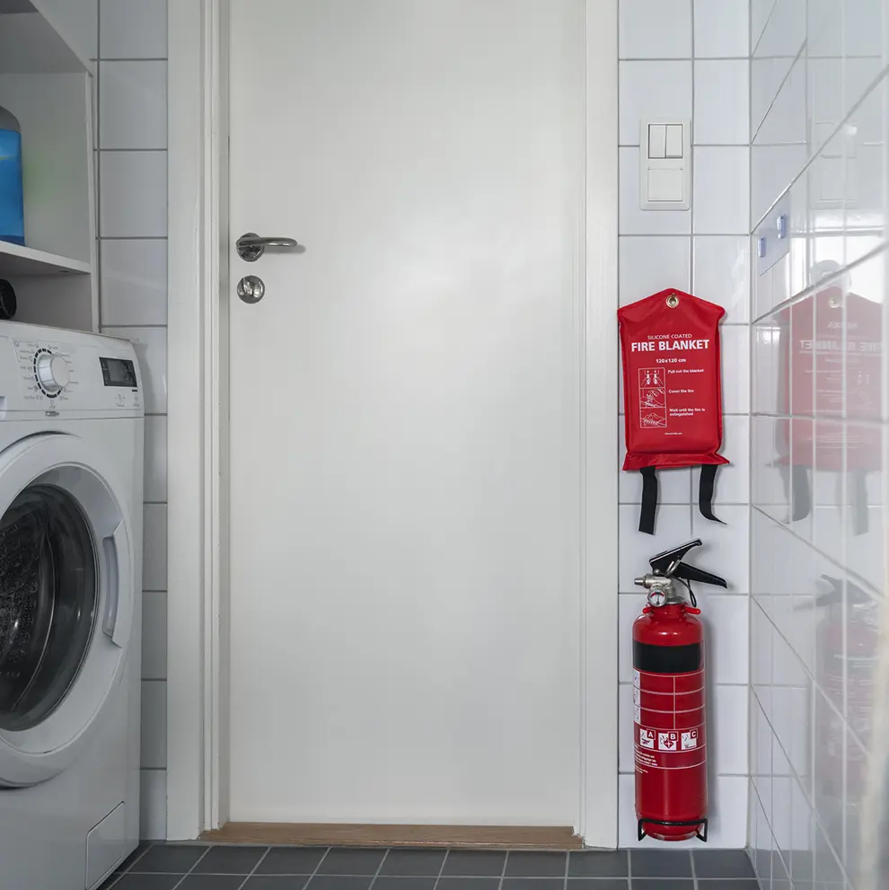 Brannslukker og brannteppe montert på vegg i et vaskerom som del av brannsikkerhet i hjemmet.