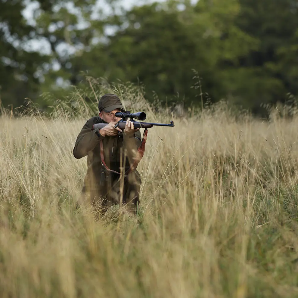 Jeger sikter med rifle i høyt gress under jakt i skogsområde.