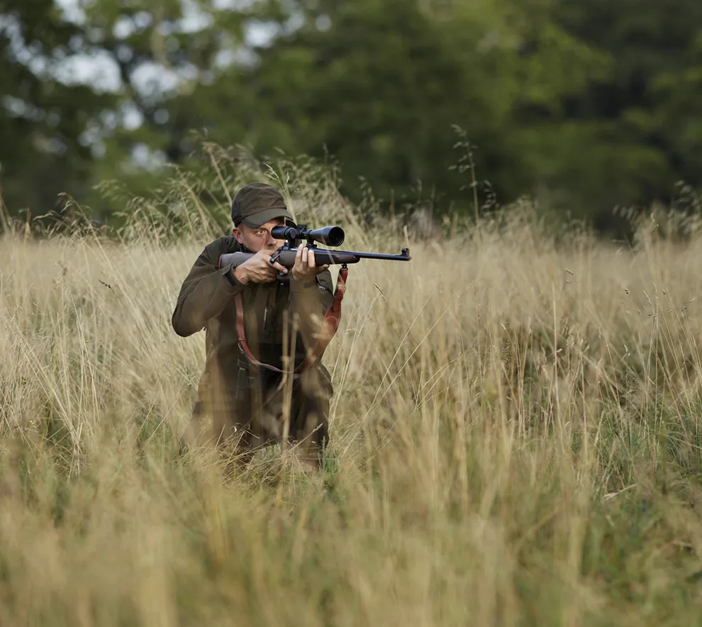 Jeger sikter med rifle i høyt gress under jakt i skogsområde.