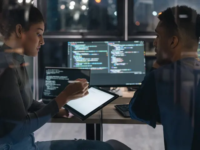 To IT-eksperter diskuterer foran skjermer med programmeringskode.