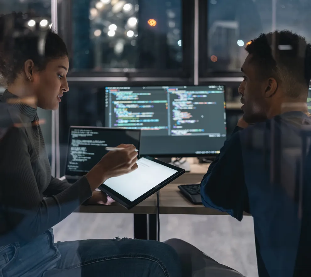To IT-eksperter diskuterer foran skjermer med programmeringskode.