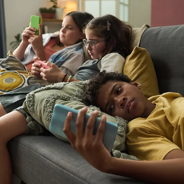 Tre barn ligger i sofaen og ser på mobiltelefoner, fokus på skjermbruk og teknologi.