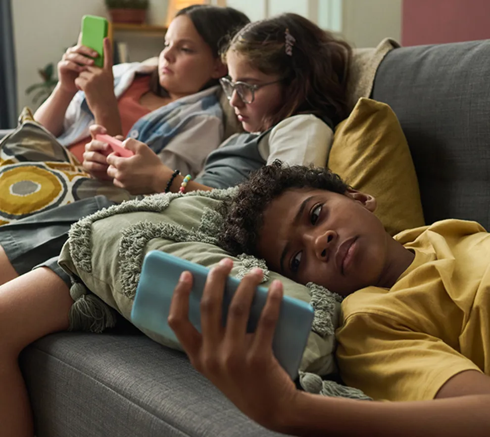 Tre barn ligger i sofaen og ser på mobiltelefoner, fokus på skjermbruk og teknologi.