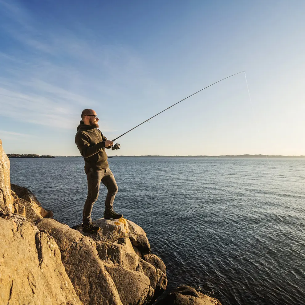 Mann fisker med fiskestang fra svaberg ved sjøen.