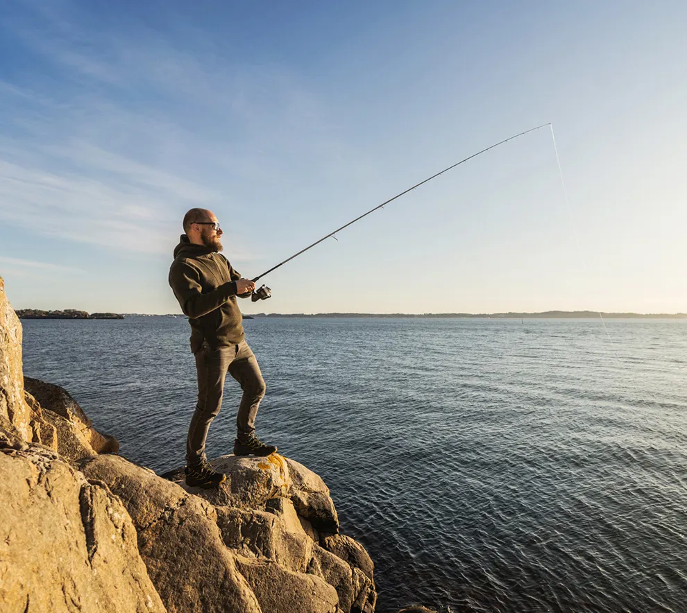 Mann fisker med fiskestang fra svaberg ved sjøen.