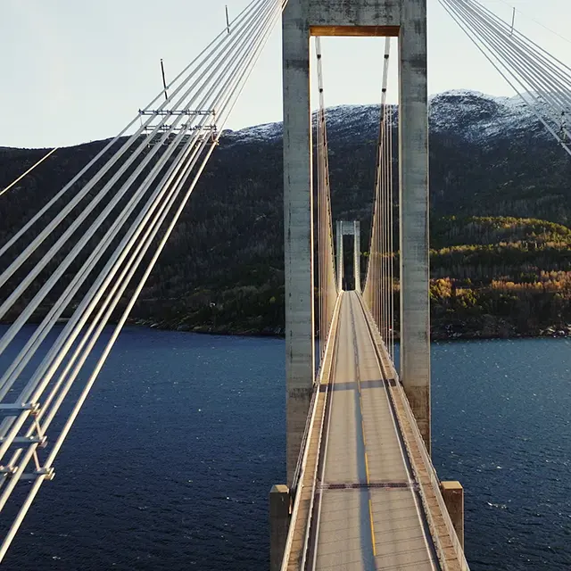 Hengebru over fjord omgitt av fjell og skog, sett fra midten av brudekket.