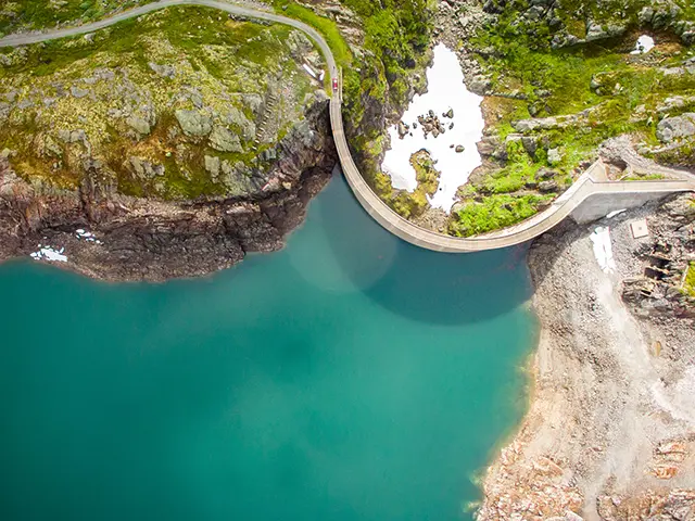 Fugleperspektiv av en turkis innskø med grønn natur og en kurvet demning opp mot vannet.