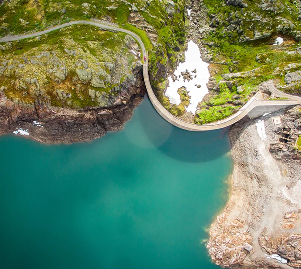 Fugleperspektiv av en turkis innskø med grønn natur og en kurvet demning opp mot vannet.
