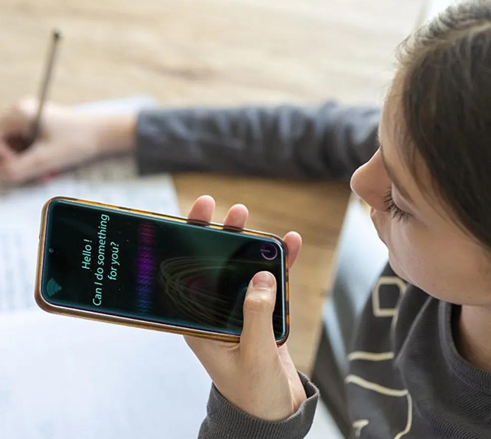 ung kvinne sitter med telefon i hånden snakker til KI, noterer samtidig